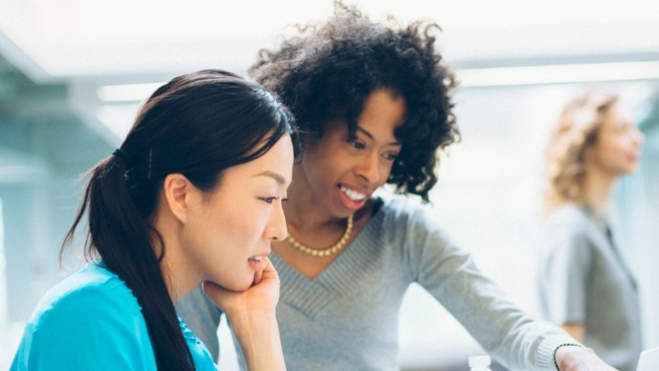 Stock photo of two women working together in an office while a group of 3 converse in the background