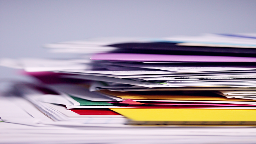 A disorganized pile of paper, folders, and envelopes, viewed from the side.